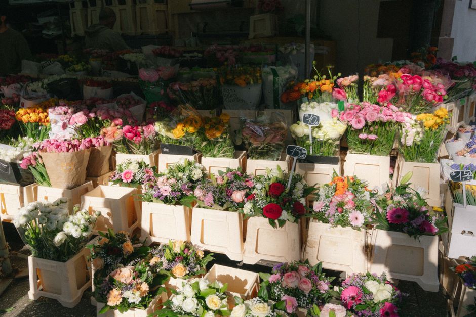A vibrant outdoor flower stall displaying a variety of fresh floral arrangements. The arrangement includes pink roses, white lilies, yellow tulips, and purple daisies, all meticulously assembled into 
