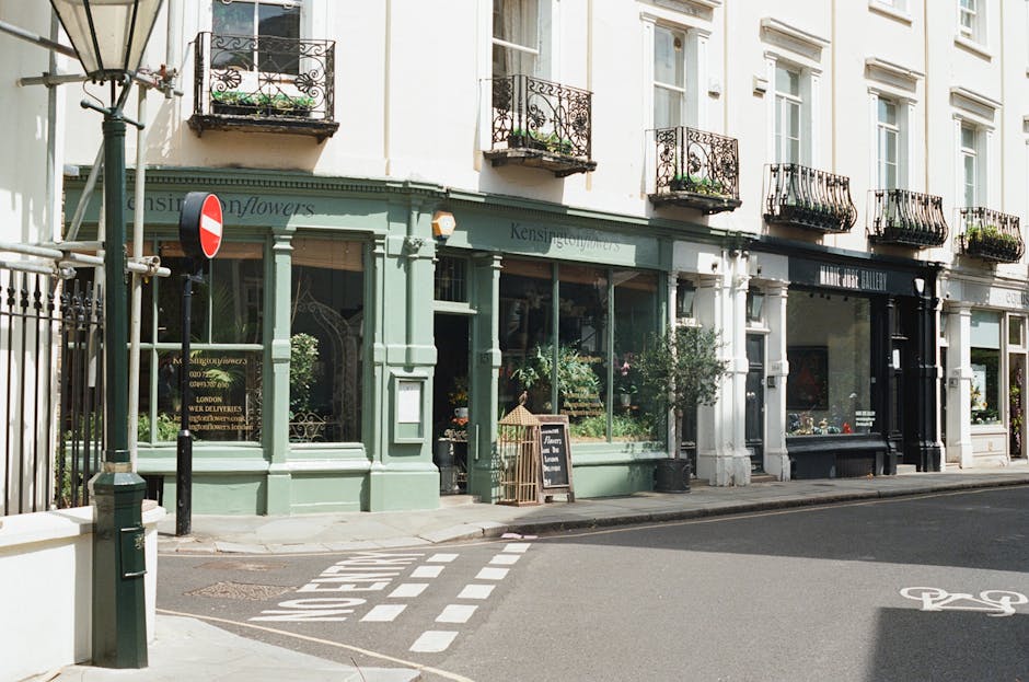 A street view showing a flower shop with a pastel green facade, featuring large glass windows displaying various potted plants and floral arrangements. The shop is situated on a corner of a quiet stre
