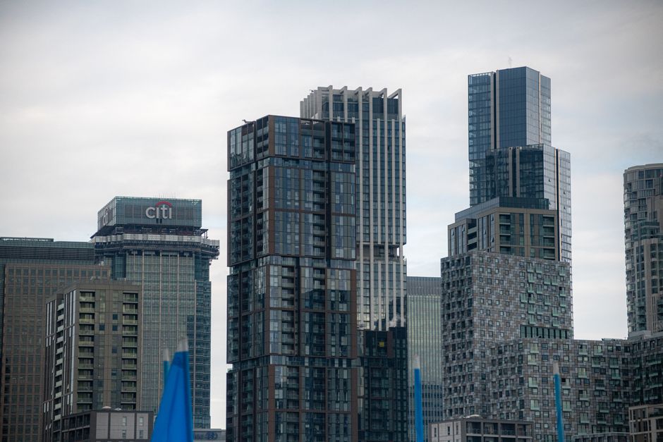A cluster of modern high-rise office buildings in Canary Wharf, London, with glass facades reflecting the cloudy sky overhead. The buildings feature various architectural styles, including blocky, geo