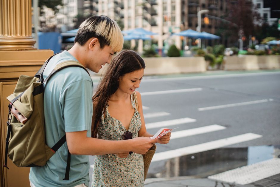 A young man and woman stand on a city sidewalk near a pedestrian crossing, engaging in a conversation while looking at a mobile phone. The young man has light blonde hair with dark roots, is wearing a