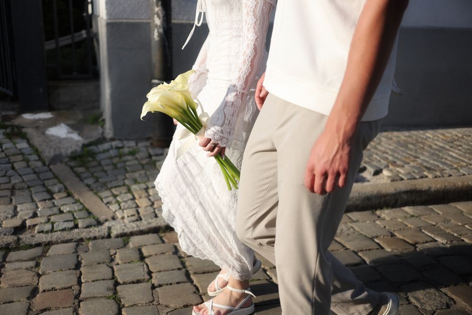 Close-up of a bride holding a bouquet of white calla lilies with long green stems, standing next to a groom dressed in beige trousers and a white shirt. The bride's white lace wedding dress is visible